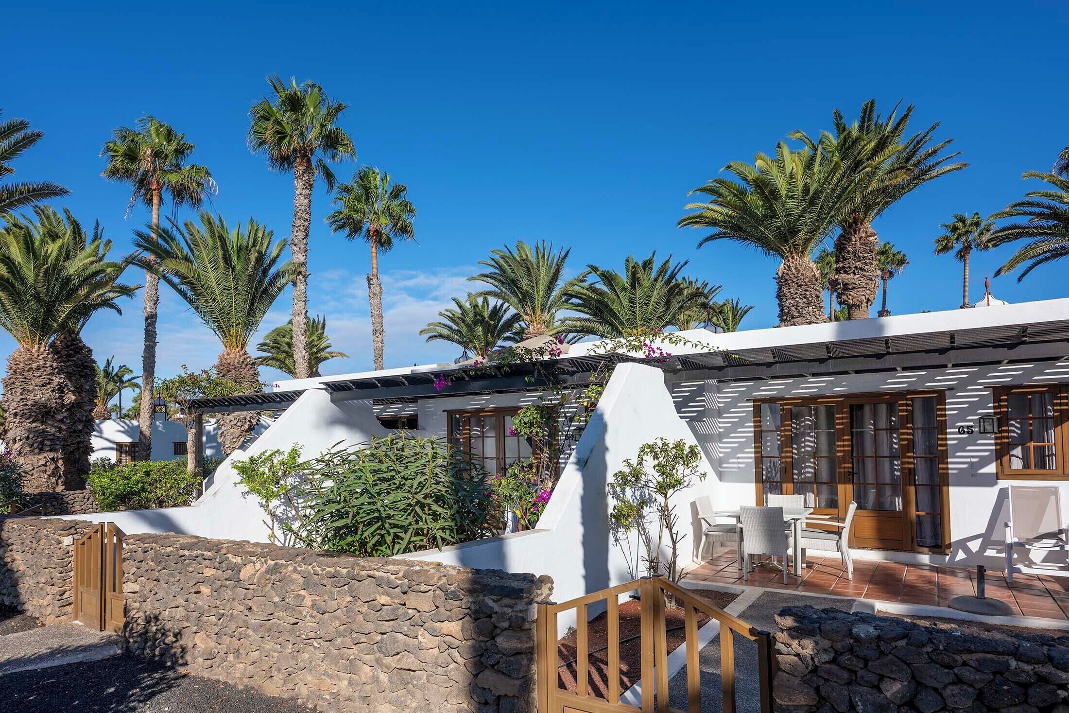 A modern villa with a stone wall entrance, a patio with outdoor furniture, and palm trees in the background.