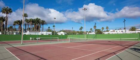 An outdoor tennis court with a net, surrounded by a green fence, palm trees, and a clear sky.