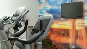 Una bicicleta estática con pantalla digital y panel de control, un televisor montado en la pared, y un mural que muestra un atardecer.