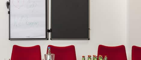 A conference room with a whiteboard displaying 'Ich - Willkommen' and a table with red chairs, water, and condiments.