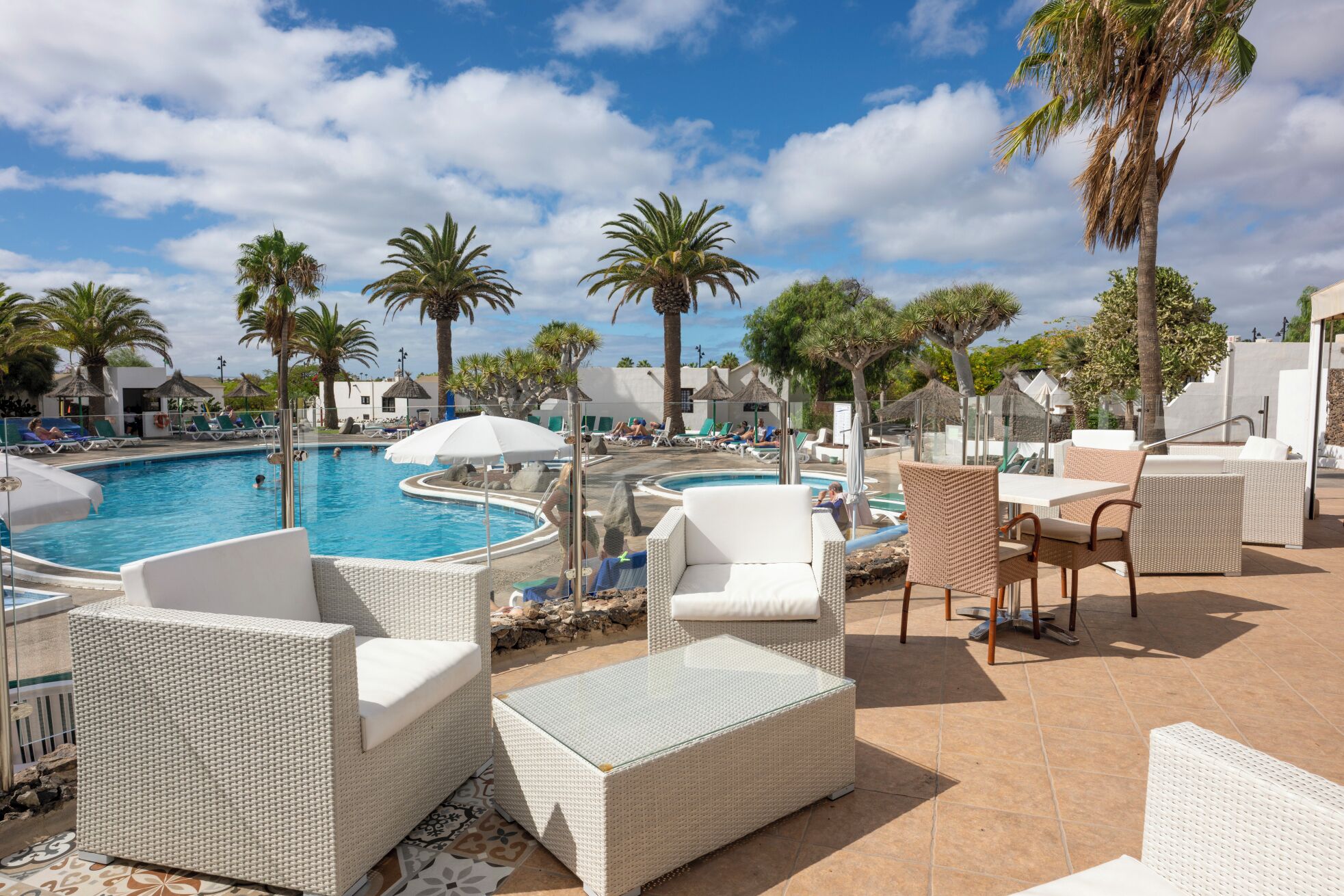 A poolside area with lounge chairs and a glass-top table, surrounded by palm trees and a clear sky.