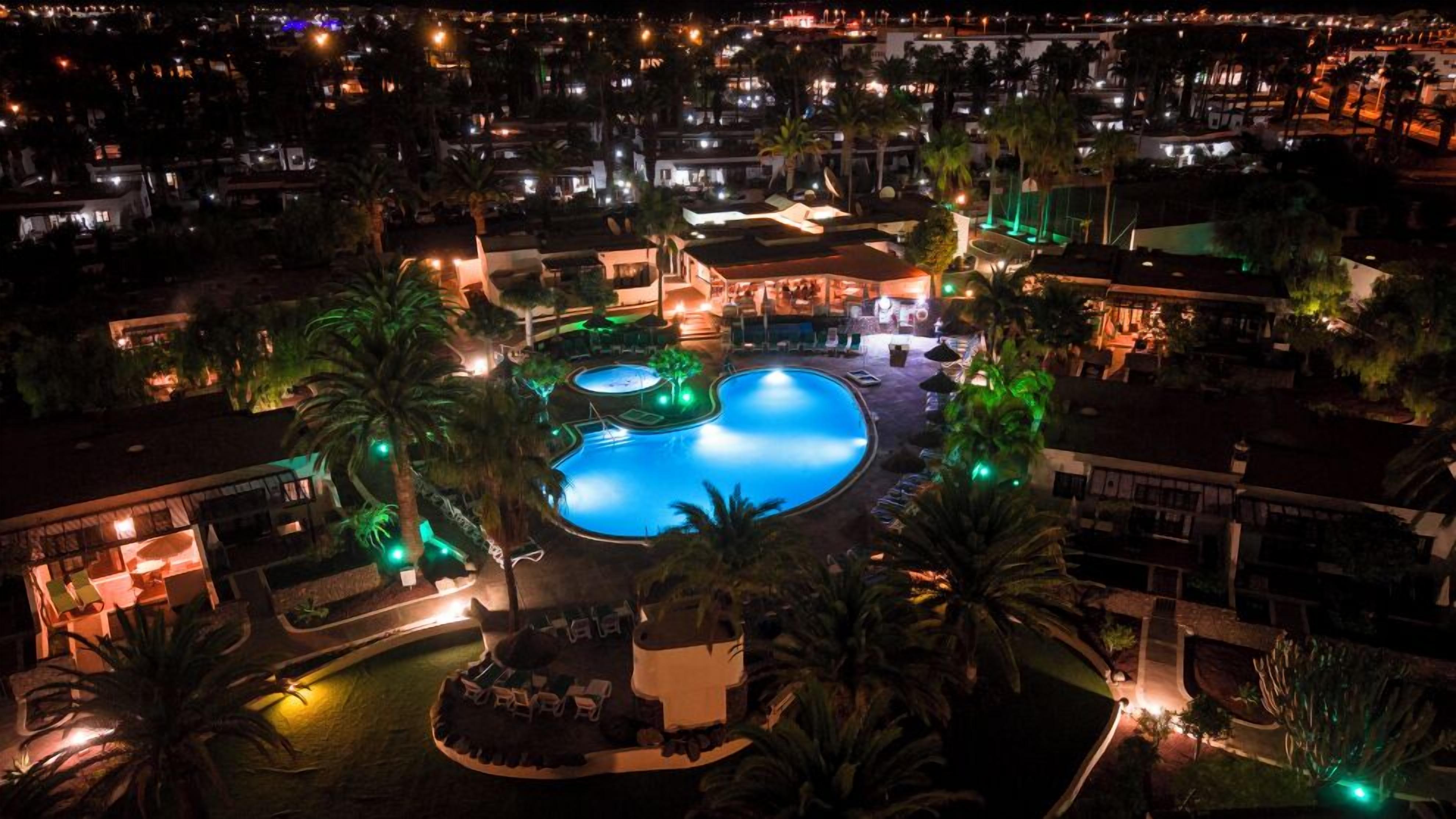 Vue nocturne d’un complexe hôtelier doté d’une grande piscine, de palmiers et d’espaces de détente extérieurs éclairés.
