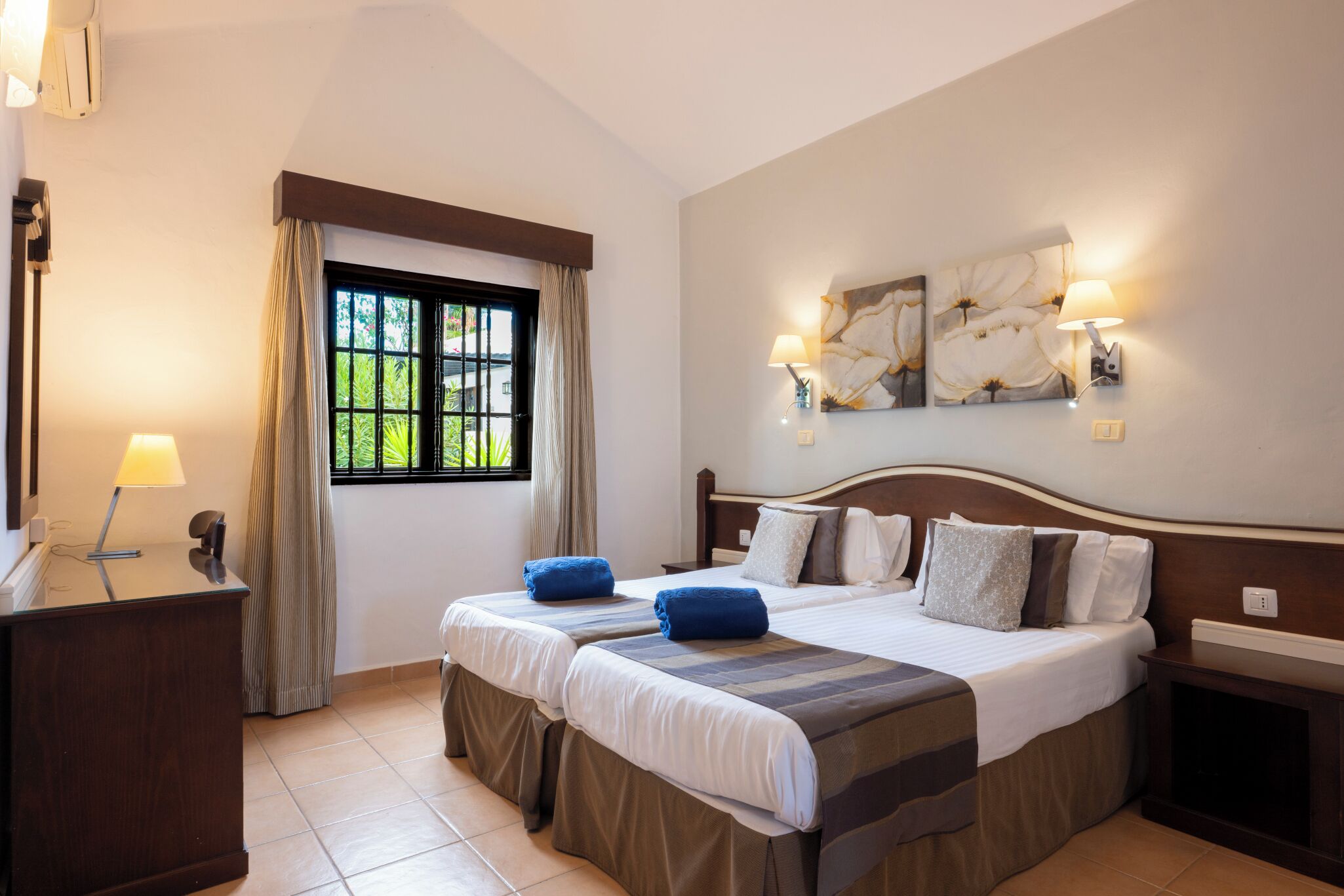 Une chambre d’hôtel avec deux lits, un bureau, une fenêtre avec des rideaux et des lampes fixées au mur.