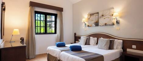 A hotel room with two beds, a desk, a window with curtains, and wall-mounted lamps.