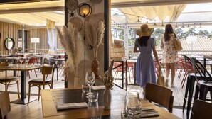 Un espace de restauration intérieur avec des tables et des chaises en bois, un vase contenant des plantes séchées et une vue sur l’espace extérieur aménagé pour les repas.