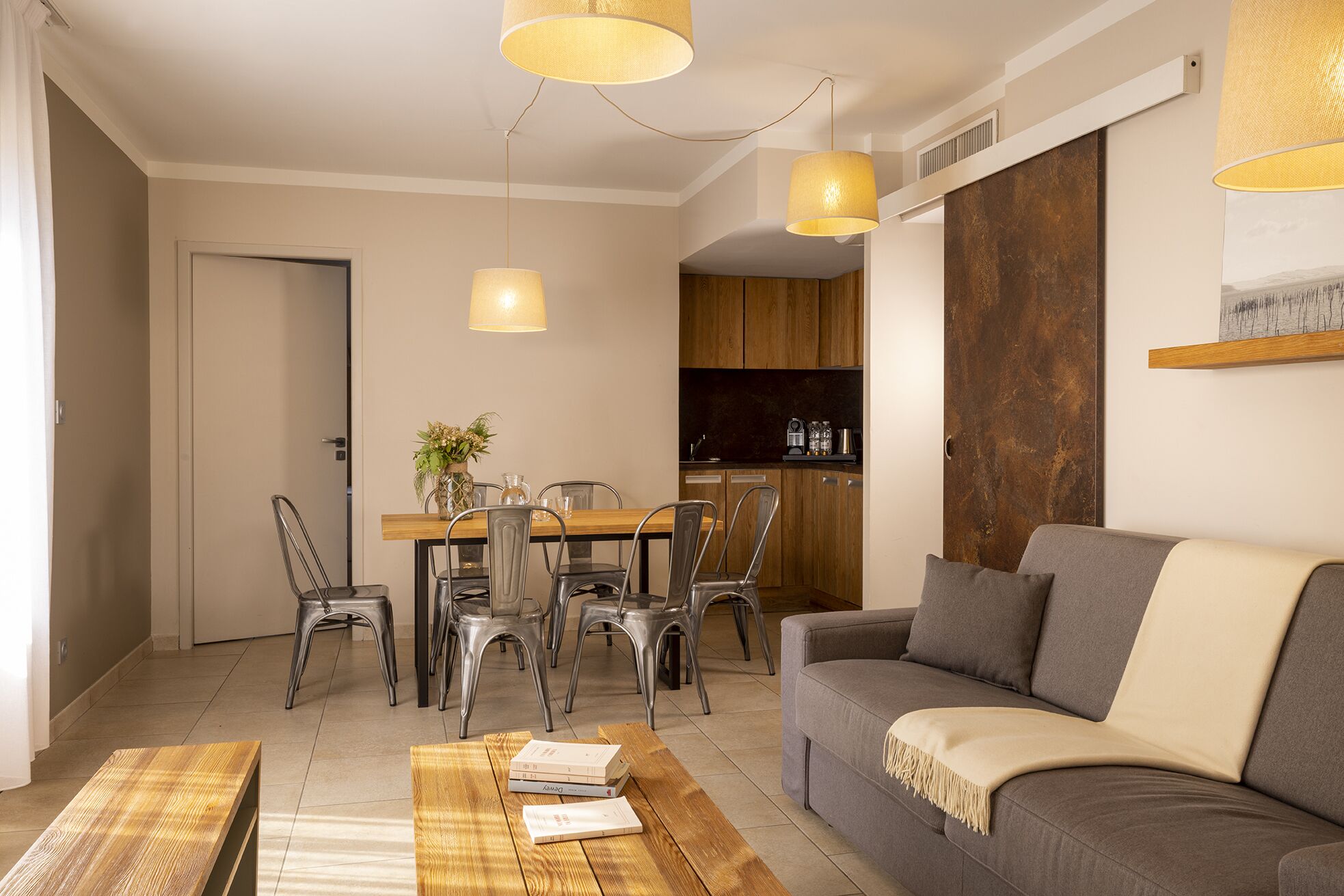A modern living room with a grey sofa, wooden coffee table, and metal chairs. A dining area with a wooden table and chairs is visible in the background.