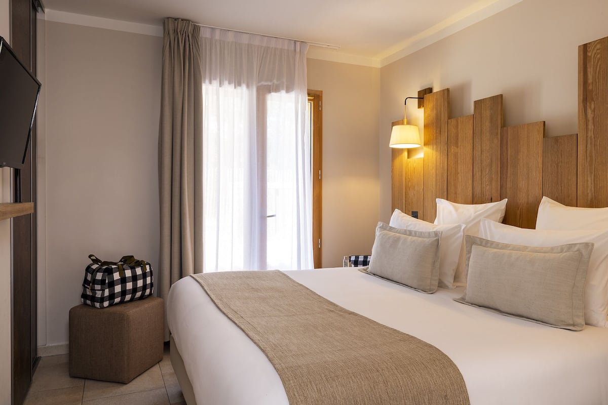 Ein Hotelzimmer mit einem großen Bett, einem Holzkopfende, einem Nachttisch, einer Lampe und einer karierten Tasche auf einem kleinen Hocker.
