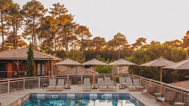 Un espace piscine doté de chaises longues et de parasols, entouré d’une terrasse en bois et de grands arbres.