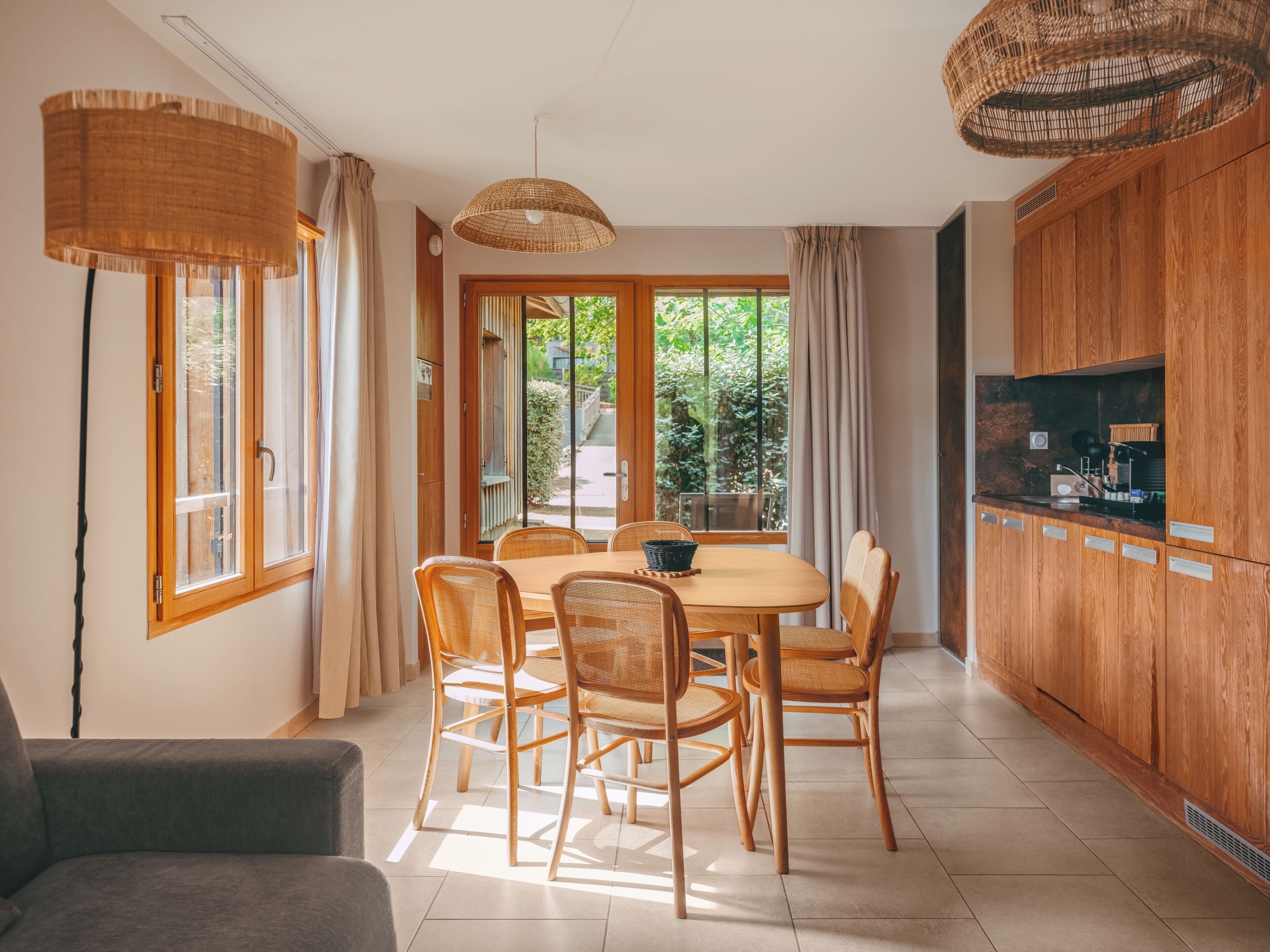 A dining area with a table and chairs, a kitchen with wooden cabinets, and a view of greenery outside.