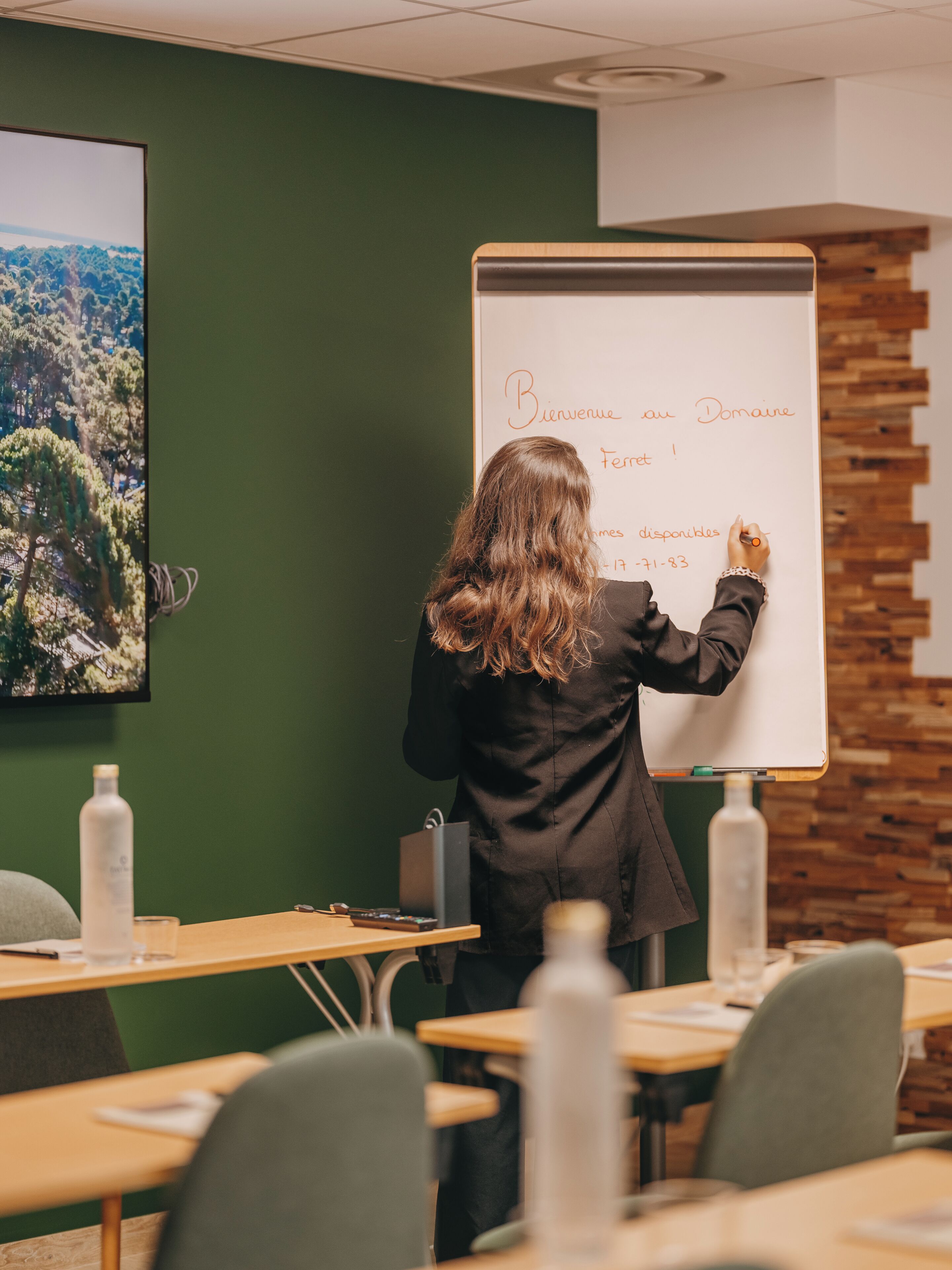 A person writing on a whiteboard in a room with green walls, wooden accents, and a desk with a laptop and water bottle.
