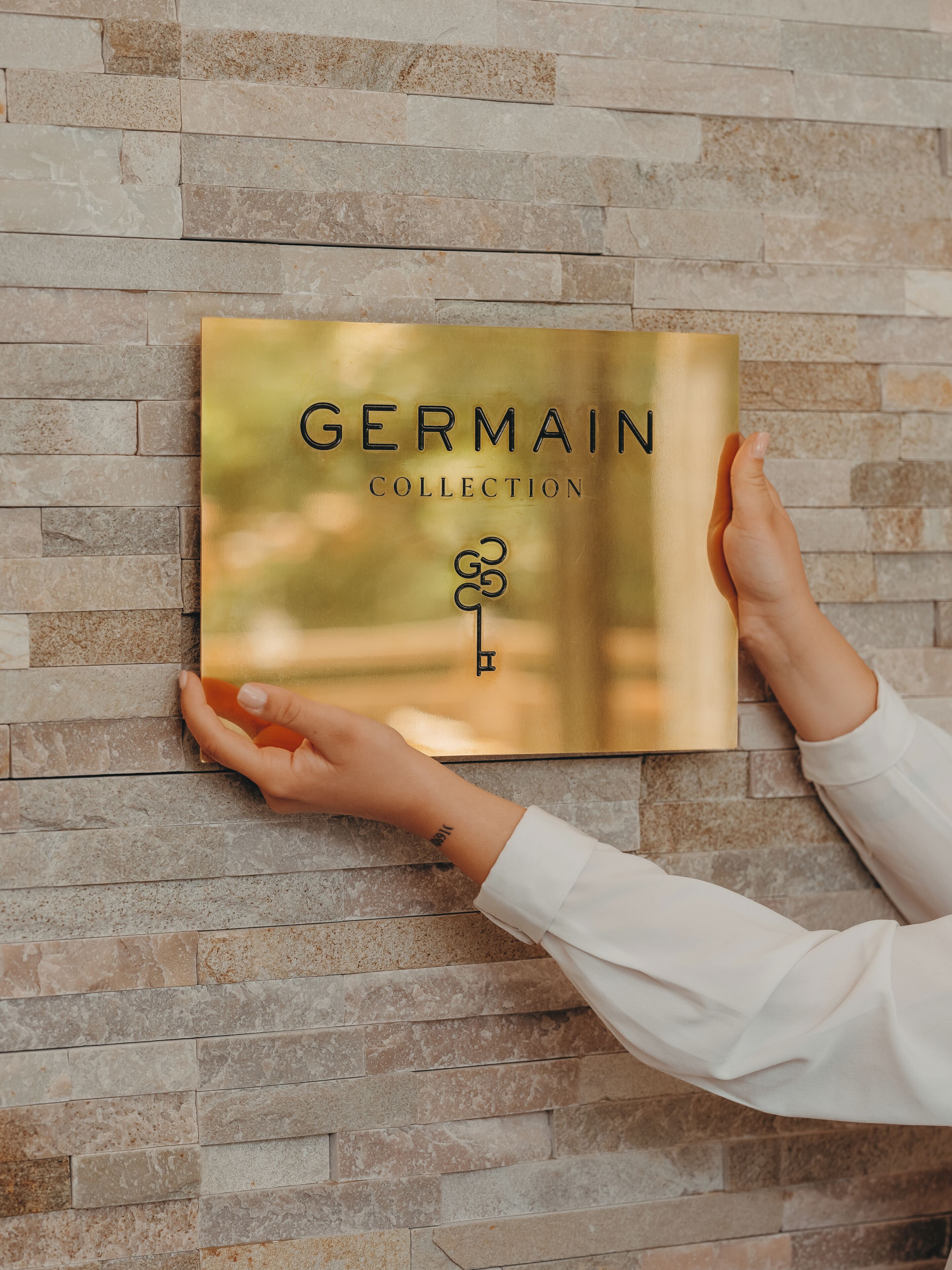 Une main tenant un objet encadré sur lequel on peut lire le texte « COLLECTION GERMAIN », le tout posé sur un mur de pierre.