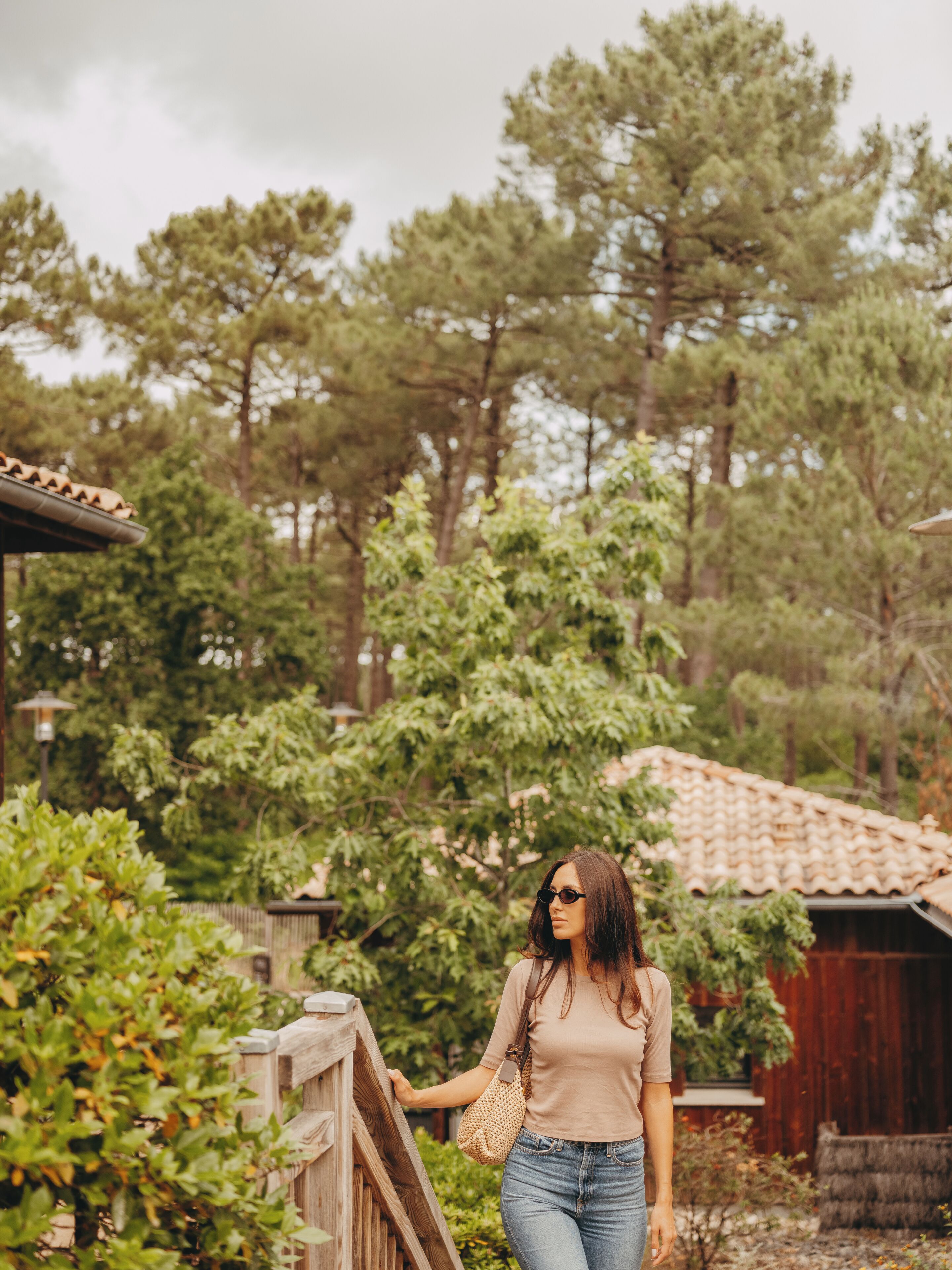 A woman walking along a wooden fence in a garden with trees and a red building in the background.