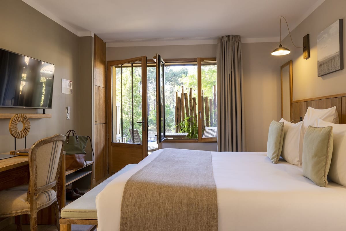 Ein Hotelzimmer mit einem großen Bett, einem Schreibtisch mit Stuhl, einem Fernseher und einem Fenster mit Blick auf Bäume.