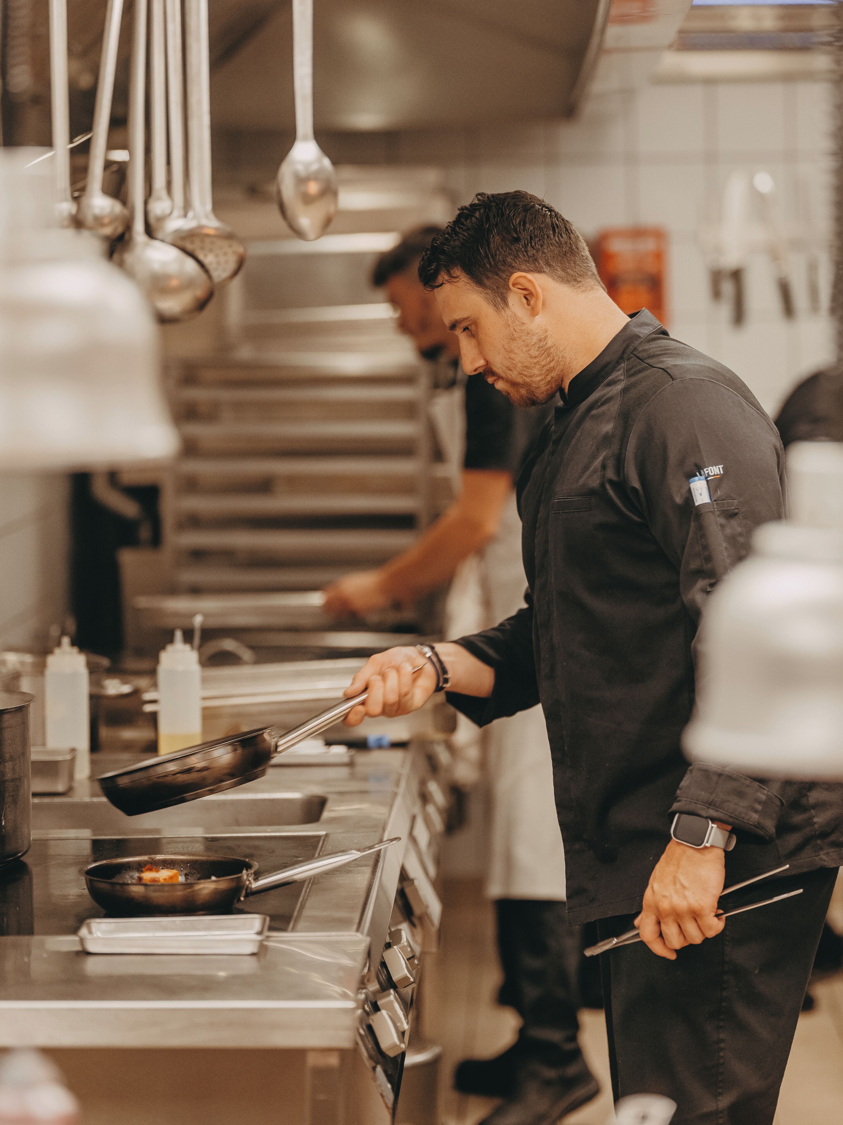 A chef in a professional kitchen with stainless steel appliances and hanging utensils.