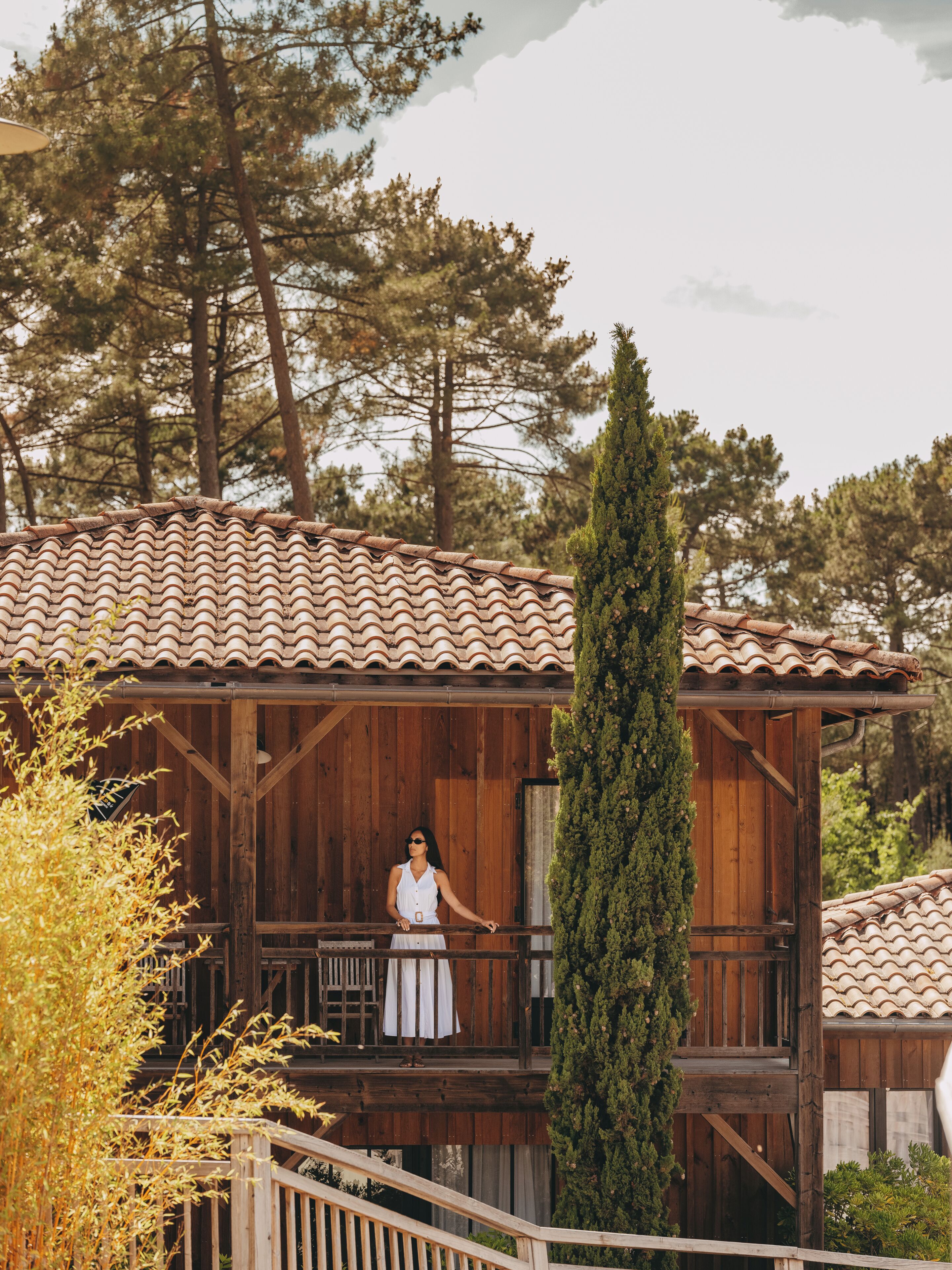 A wooden cabin with a tiled roof, a balcony, and a tall evergreen tree.