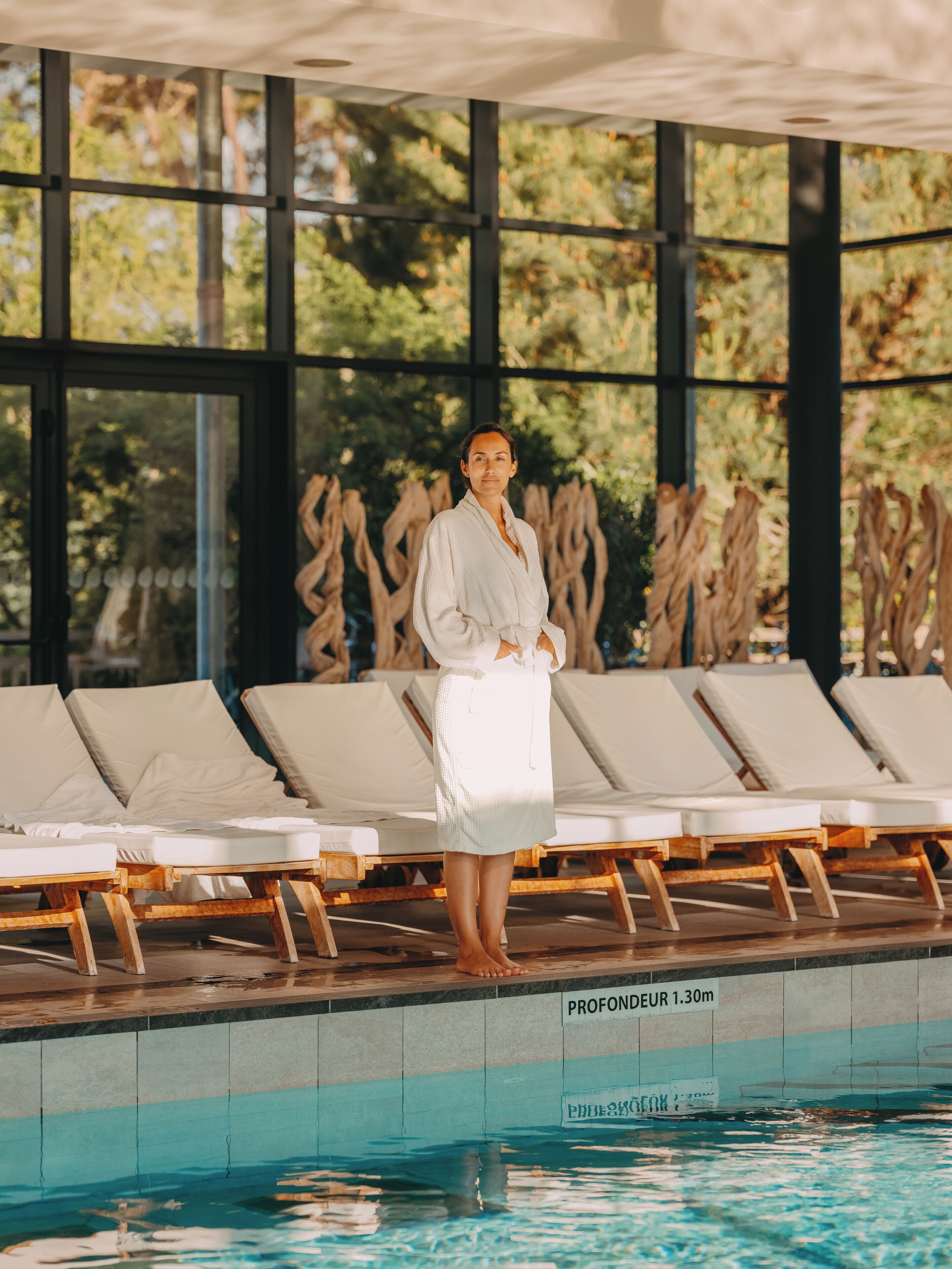 Zona de piscina con sillones de descanso, una persona con bata y un cartel que indica una profundidad de 1,30 metros.