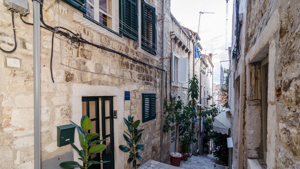 A narrow stone alleyway with potted plants and a door with a sign. - Apartments Raic (Dubrovnik)