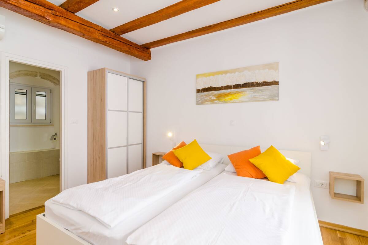 A bedroom with a bed, wooden beams, a painting, and a door leading to a bathroom.