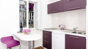 A modern kitchen with white and purple cabinets, a white countertop, a stainless steel sink, and a window with a view of a cow and flowers. - Apartments Raic (Dubrovnik)