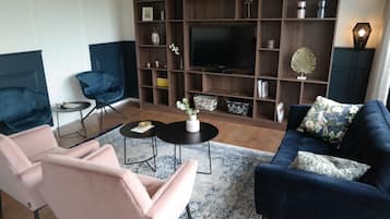 Ein modernes Wohnzimmer mit einem dunkelblauen Sofa, einem rosa Sessel und einem blauen Sessel. Ein Flachbildfernseher ist an einer Holzwandkonsole befestigt.