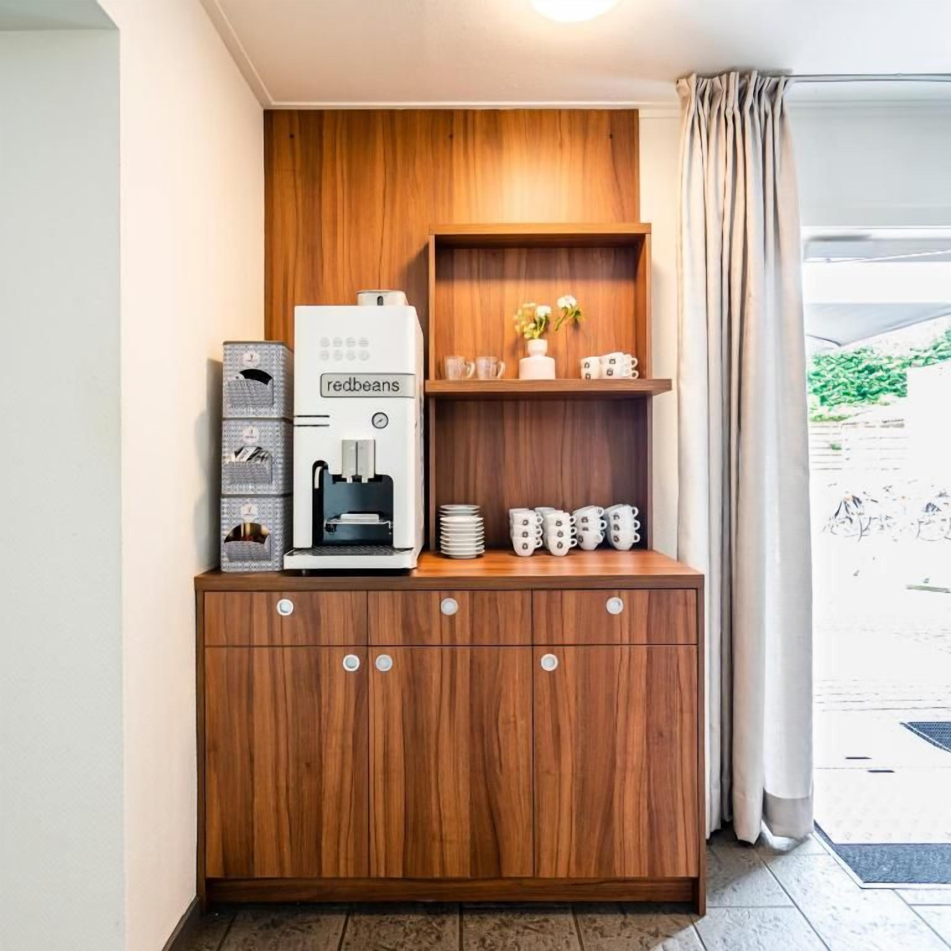 A compact kitchenette with a coffee machine, a small shelf with decorative items, and a view of the outdoors through a window.