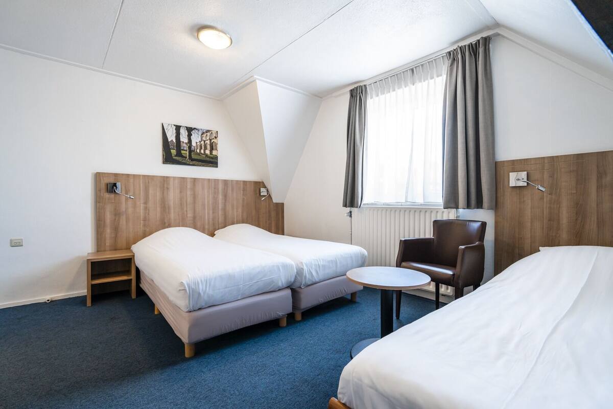Et hotelværelse med to senge, et lille bord, en stol, et billede på væggen og et vindue med gardiner.
