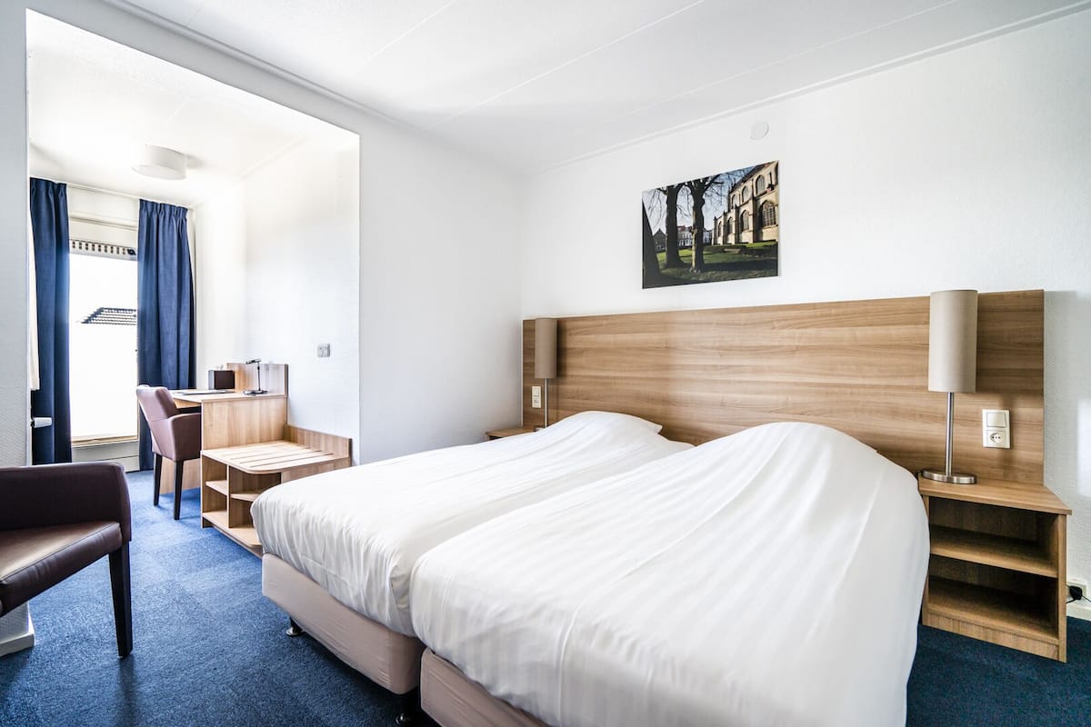 Et hotelværelse med to senge, et skrivebord, en stol, en lampe og et billede på væggen.