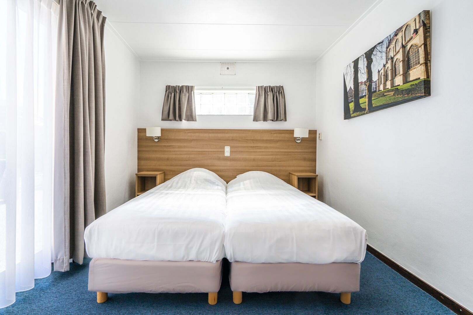 Una cama doble con cabecera de madera, dos mesitas de noche, una ventana con cortinas y un cuadro en la pared.