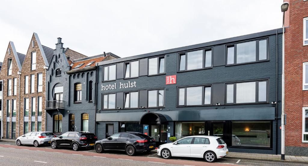 Een hotel genaamd 'hotel hulst' met een moderne gevel, geparkeerde auto's ervoor en aangrenzende traditionele gebouwen.