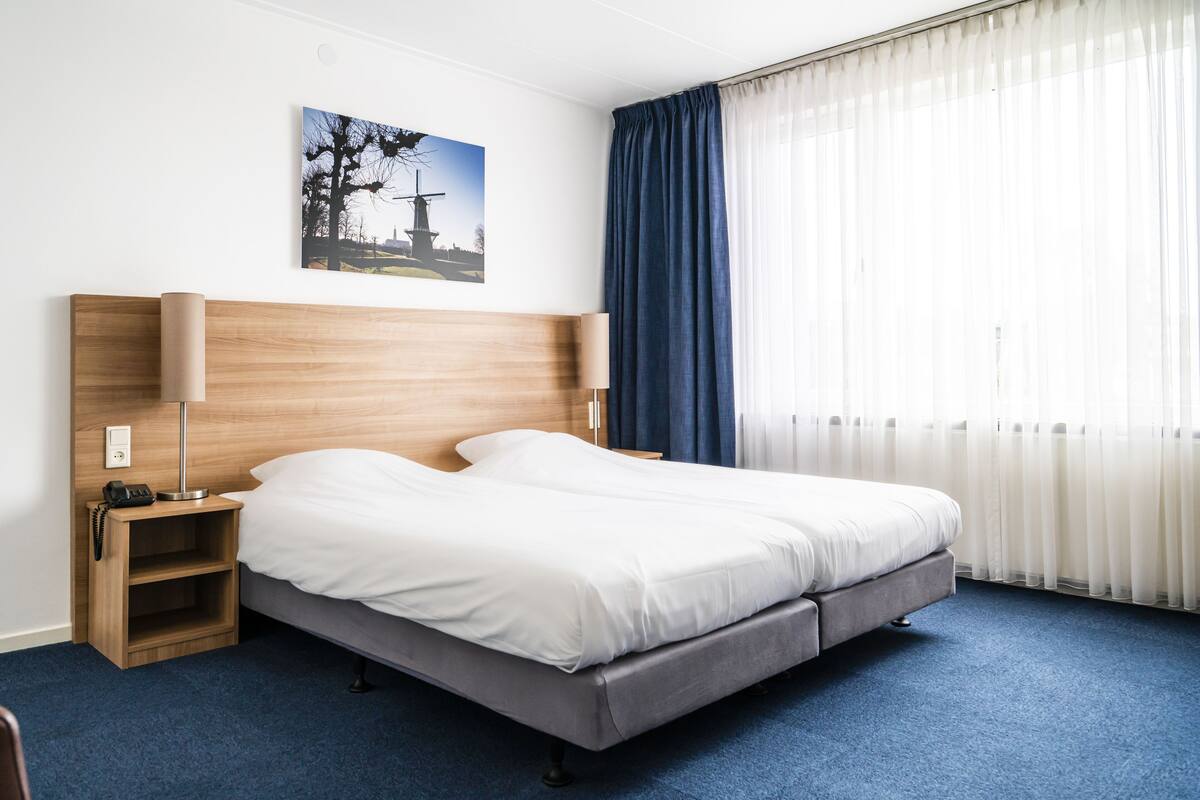 Et hotelværelse med en dobbeltseng, et natbord med lampe, en træsenggavl, et blågulvstæppe, et vindue med lette gardiner og et billede på væggen.