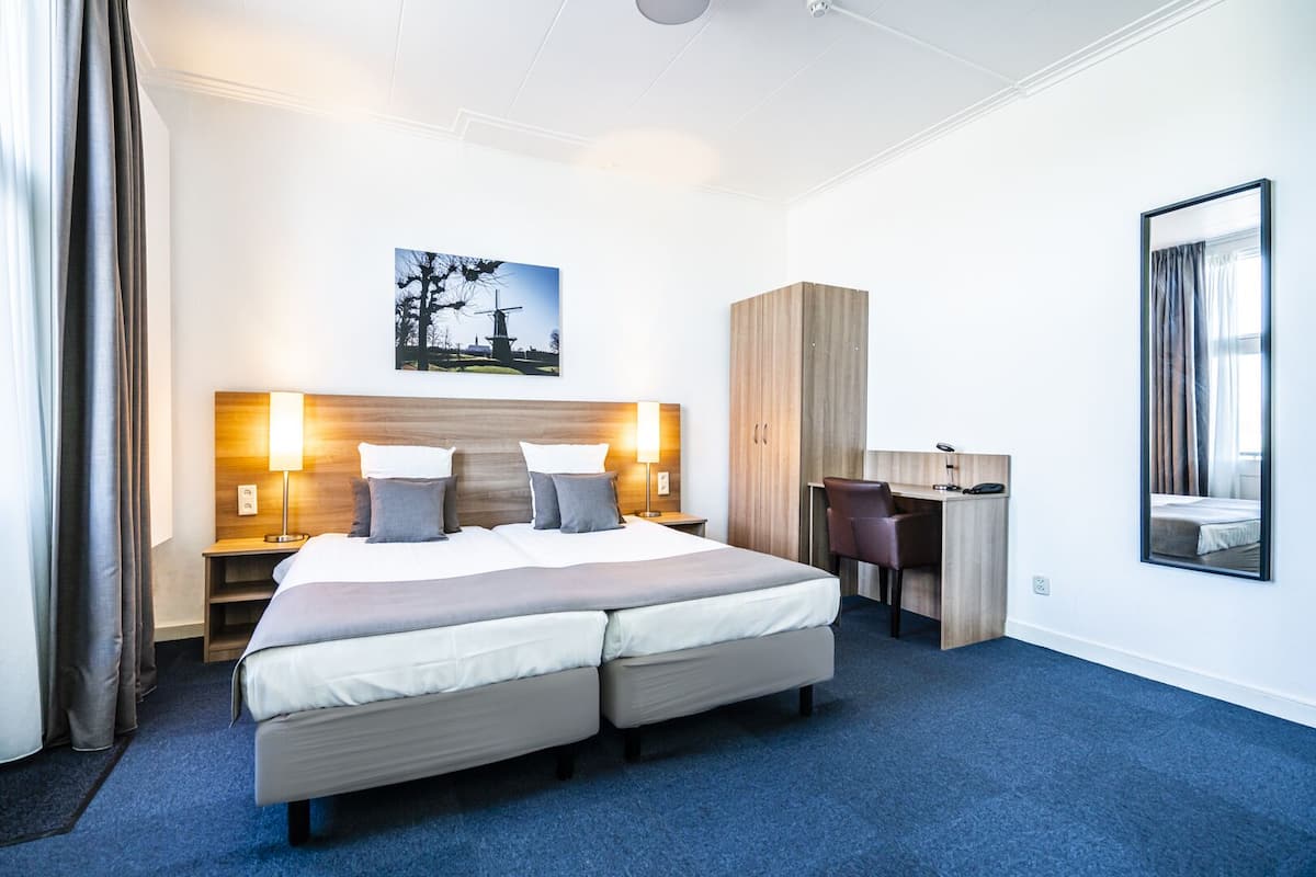 Et hotelværelse med en stor seng, to sengeborde med lamper, et skrivebord, et garderobeskab og et billede på væggen.