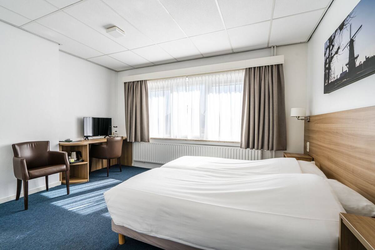 Et hotelværelse med en stor seng, et skrivebord med stol, et fjernsyn og et vindue med gardiner.