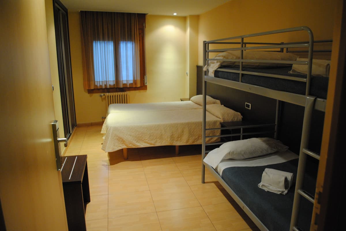 A dormitory room with bunk beds, a window with curtains, a radiator, and a wooden floor.