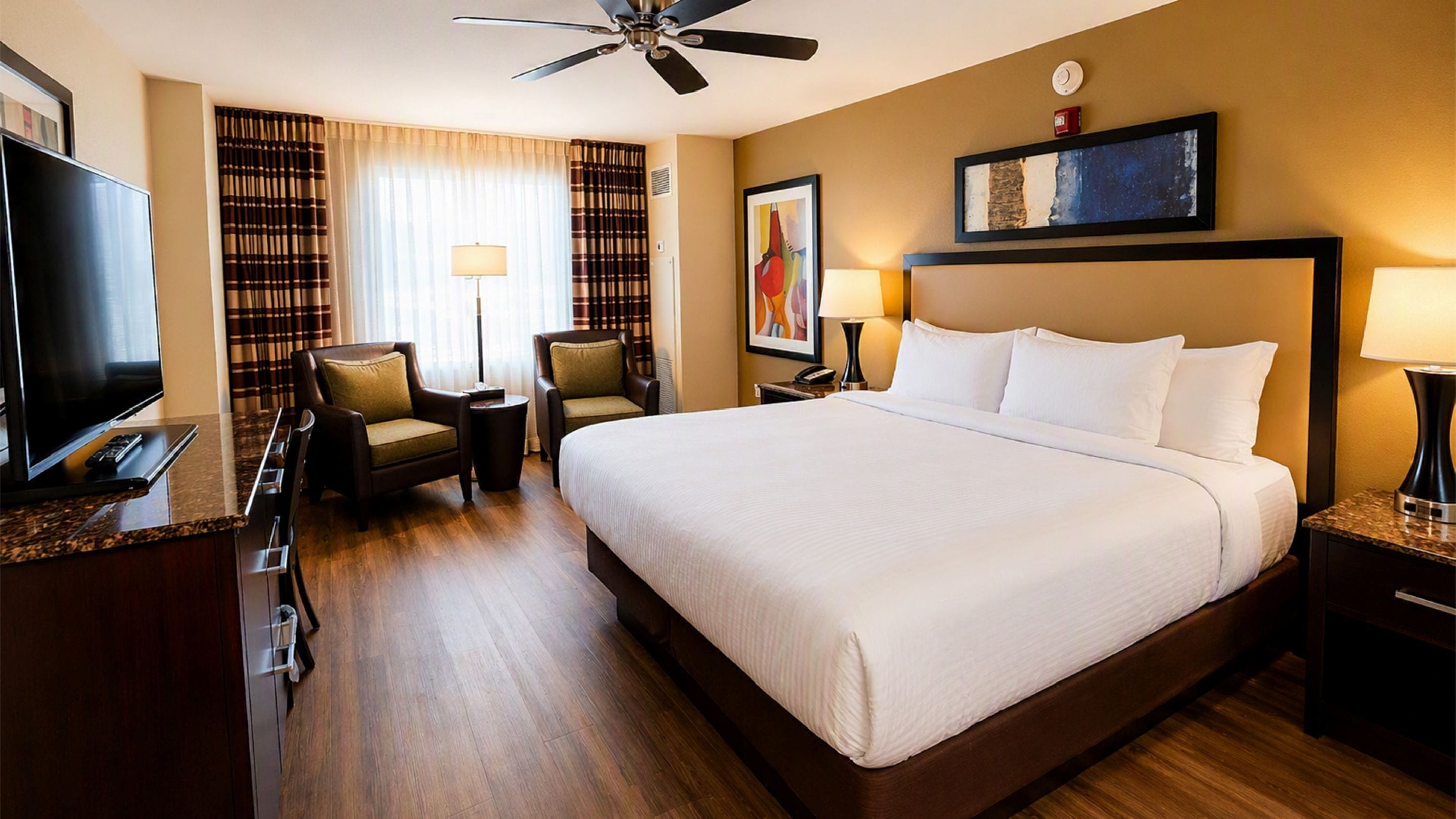 A hotel room with a large bed, a television, a desk with a computer, a chair, and a ceiling fan.