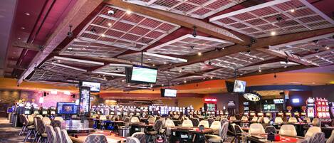 Un casino con múltiples mesas de juego y sillas, una pantalla grande mostrando juegos, y un techo con vigas a la vista y luminarias.