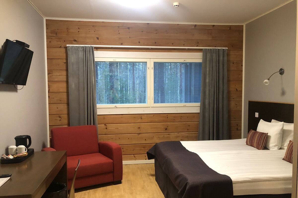Et hotelværelse med trævæg, seng med hvide sengetøj, en rød lænestol, skrivebord med kaffemaskine og et vindue med persienner.