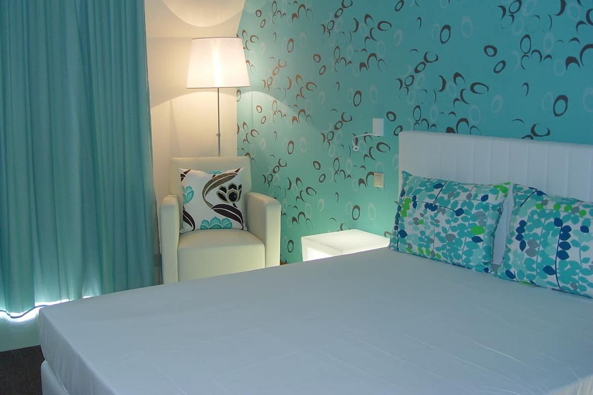 A hotel room with a bed, a chair, a nightstand, and a wall with a pattern.
