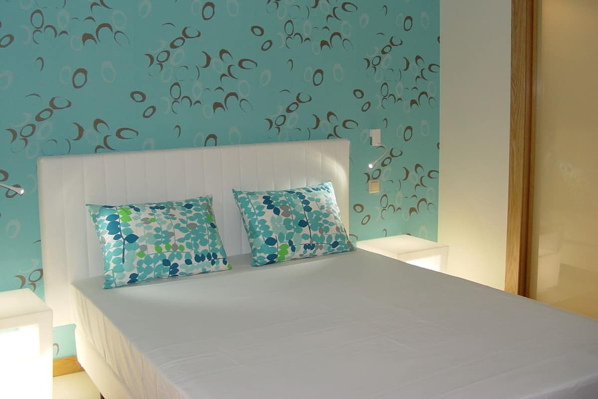 A bed with a white headboard and two decorative pillows, a nightstand with a lamp, and a wall with a repeating pattern.