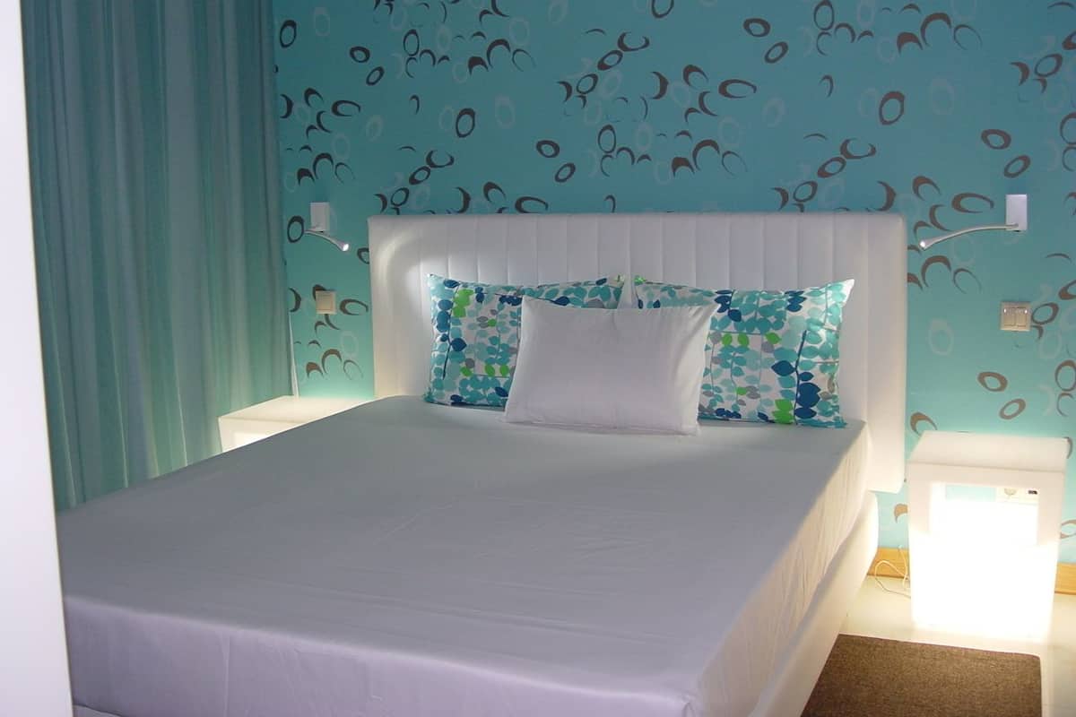 A neatly made bed with a white headboard and pillows, a light blue patterned wall, and a white nightstand with a lamp.