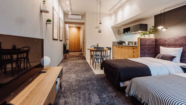 Une chambre d’hôtel moderne équipée d’un lit, d’une table de chevet et d’un petit bureau avec une chaise. On y trouve également un coin cuisine avec des placards et un four à micro-ondes.
