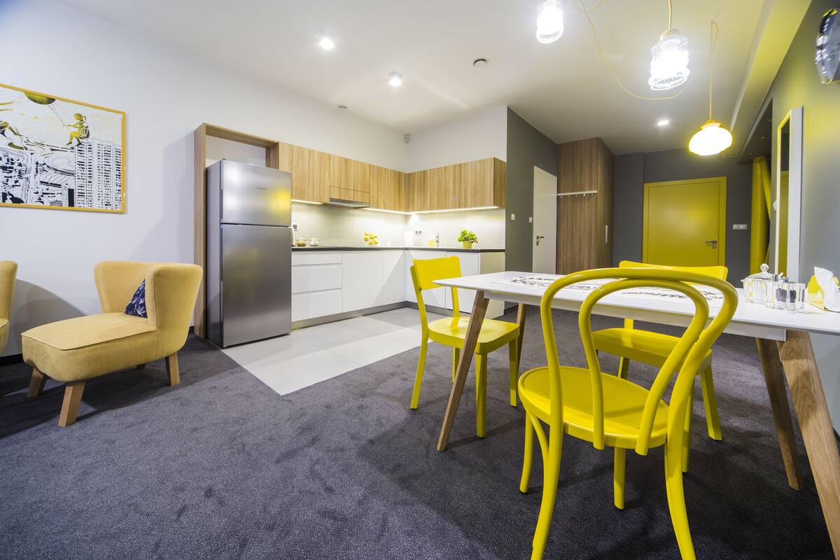 A modern kitchen with a refrigerator, wooden cabinets, and a dining area with yellow chairs and a white table.