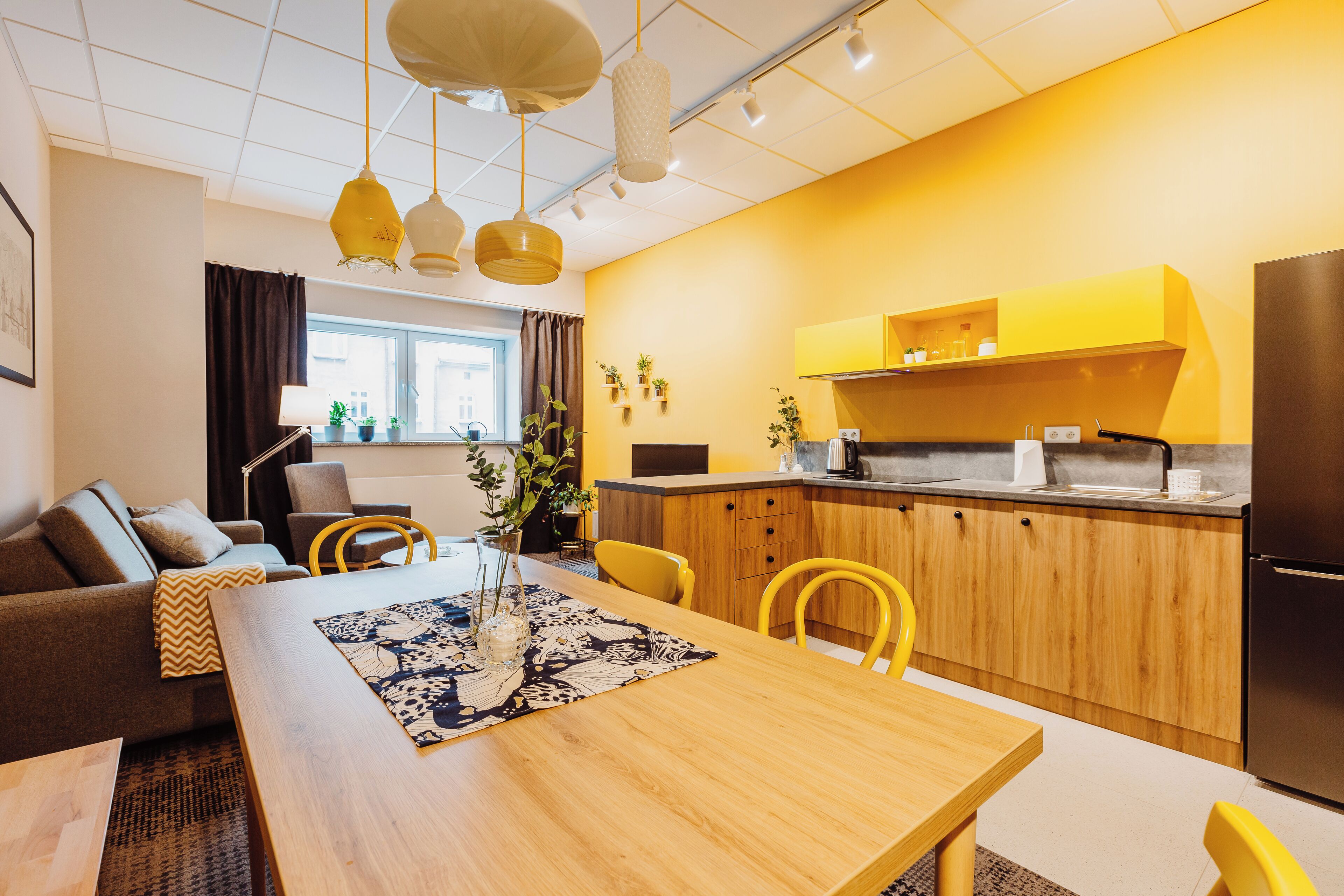 A modern kitchen with wooden cabinets, a built-in oven, and a refrigerator. There is a dining area with a wooden table and yellow chairs.