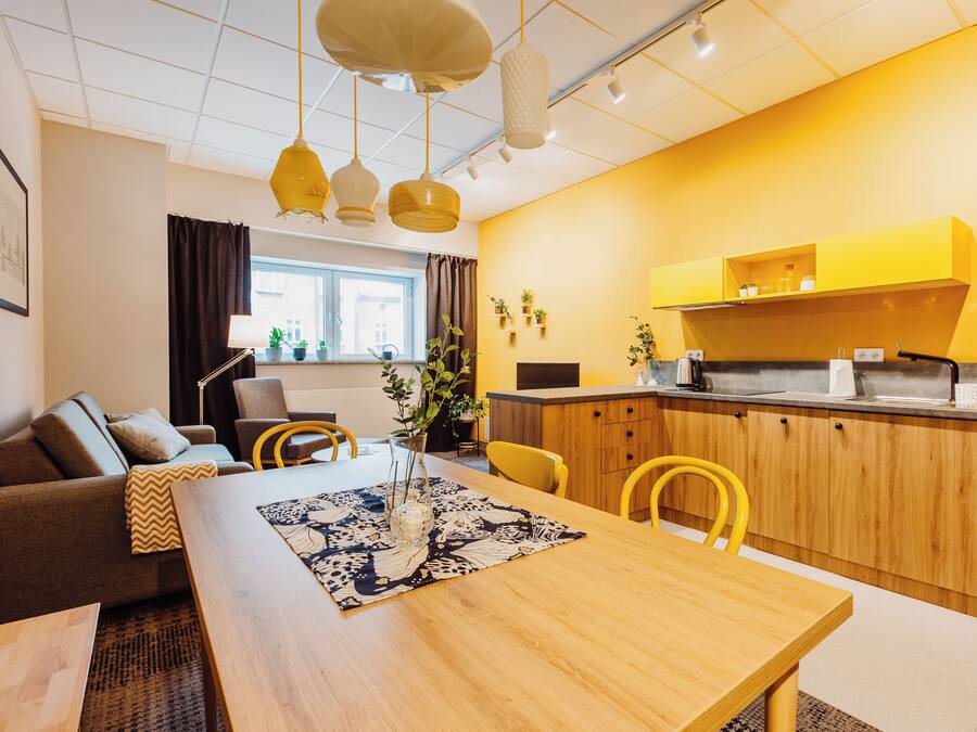 A modern kitchen with wooden cabinets, a built-in oven, and a refrigerator. There is a dining area with a wooden table and yellow chairs.