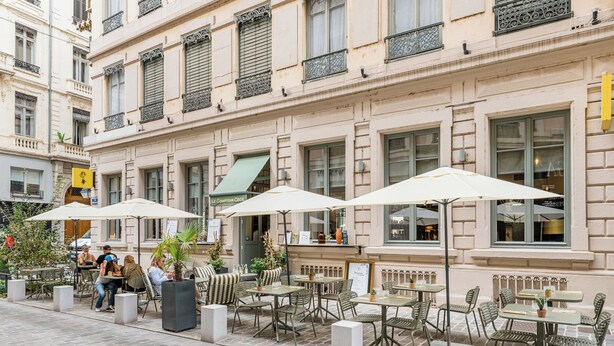 Un café al aire libre con mesas y sillas, sombrillas y una fachada de edificio con ventanas y balcones.