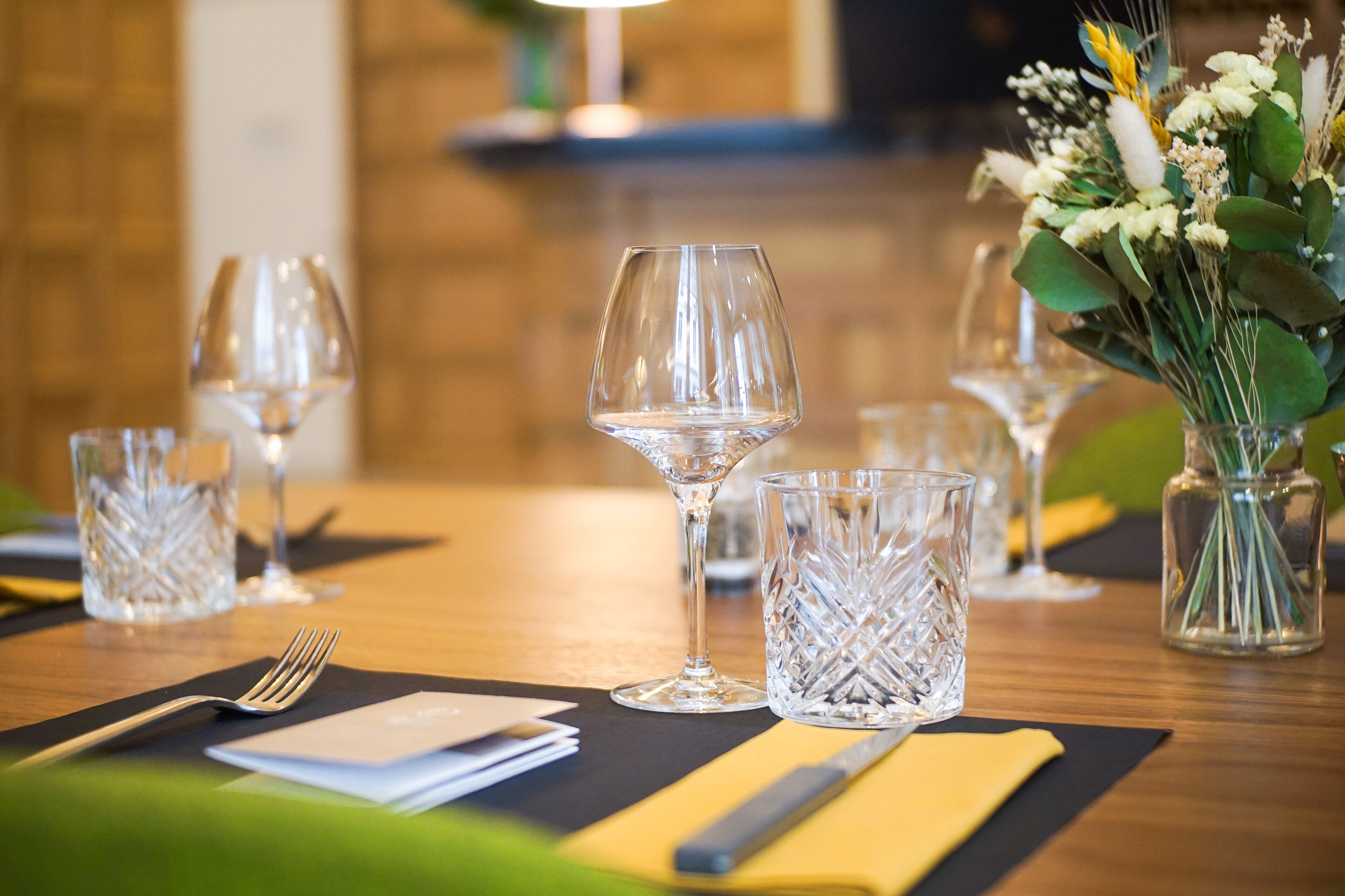 A dining table set with wine glasses, cut crystal glasses, a vase with flowers, and a fork on a black placemat.