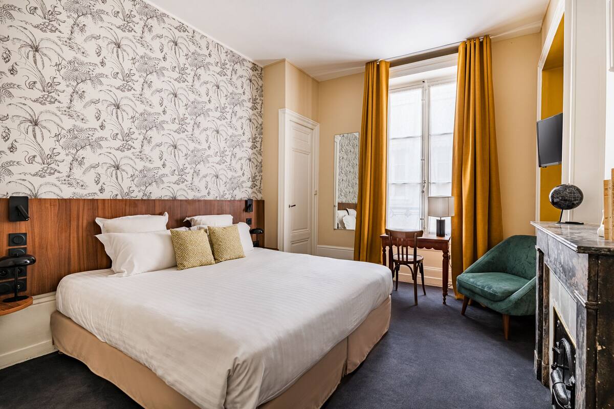A hotel room with a large bed, a desk, a chair, a television, and a fireplace.