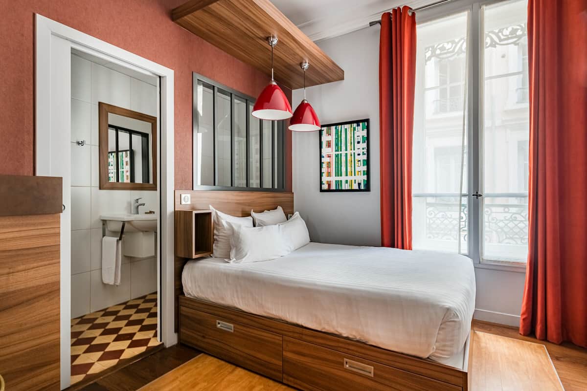 A bedroom with a wooden bed, a white bedside table, a mirror, a window with red curtains, and a view of buildings outside.