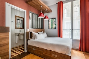 A bedroom with a wooden bed, a white bedside table, a mirror, a window with red curtains, and a view of buildings outside. - Globe et Cecil Hôtel (Lyon)