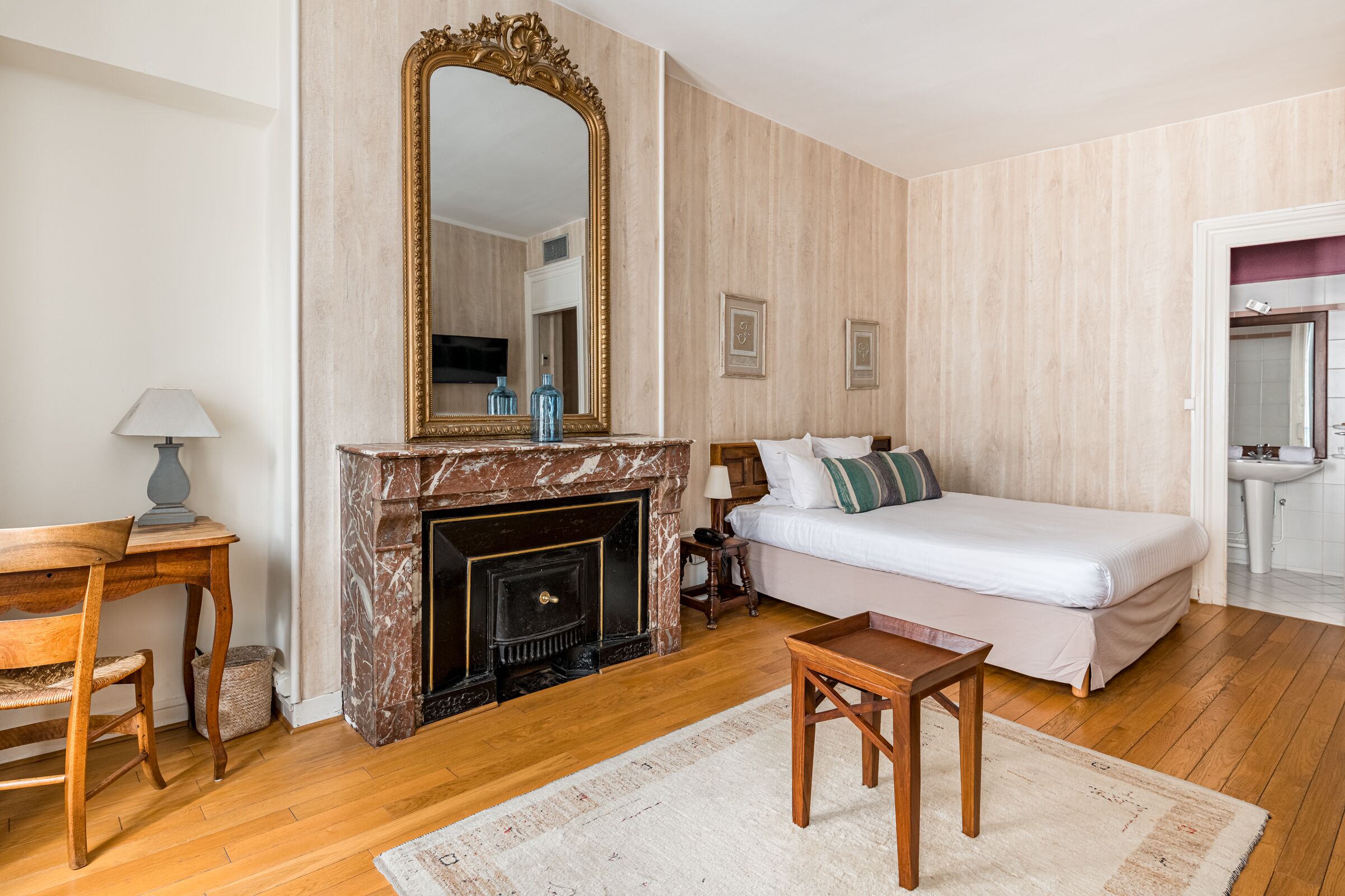 Une chambre avec un grand miroir, une cheminée, un lit avec une literie blanche, une table en bois et un petit tapis.