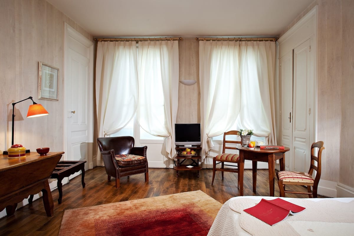 A hotel room with a wooden floor, a bed, a desk with a lamp, a television, and two chairs.